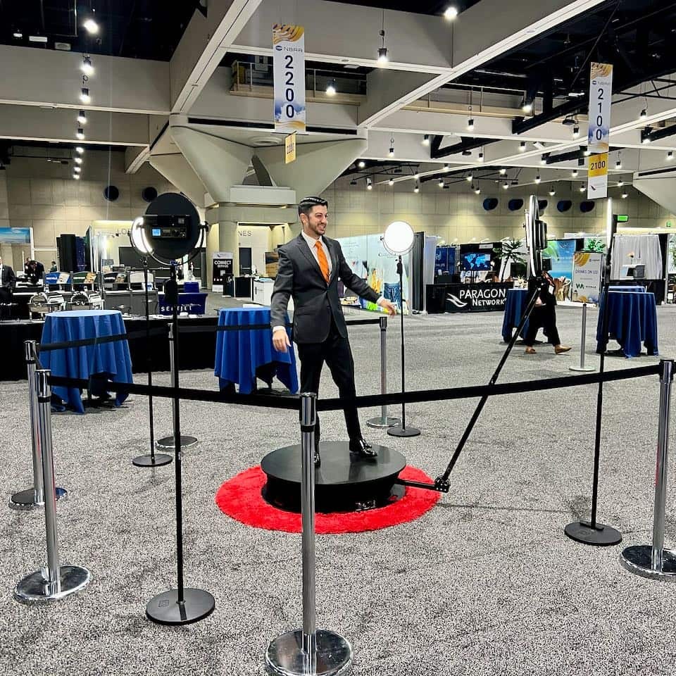 A man in a suit and orange tie stands on the platform inside a ring of stanchions on a convention center floor with exhibitor booths behind him