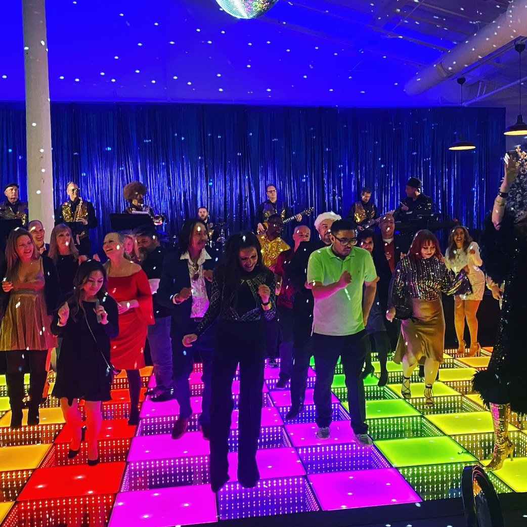 A crowd dances on a multicolor checkerboard LED floor in front of a band and a blue starlight drape