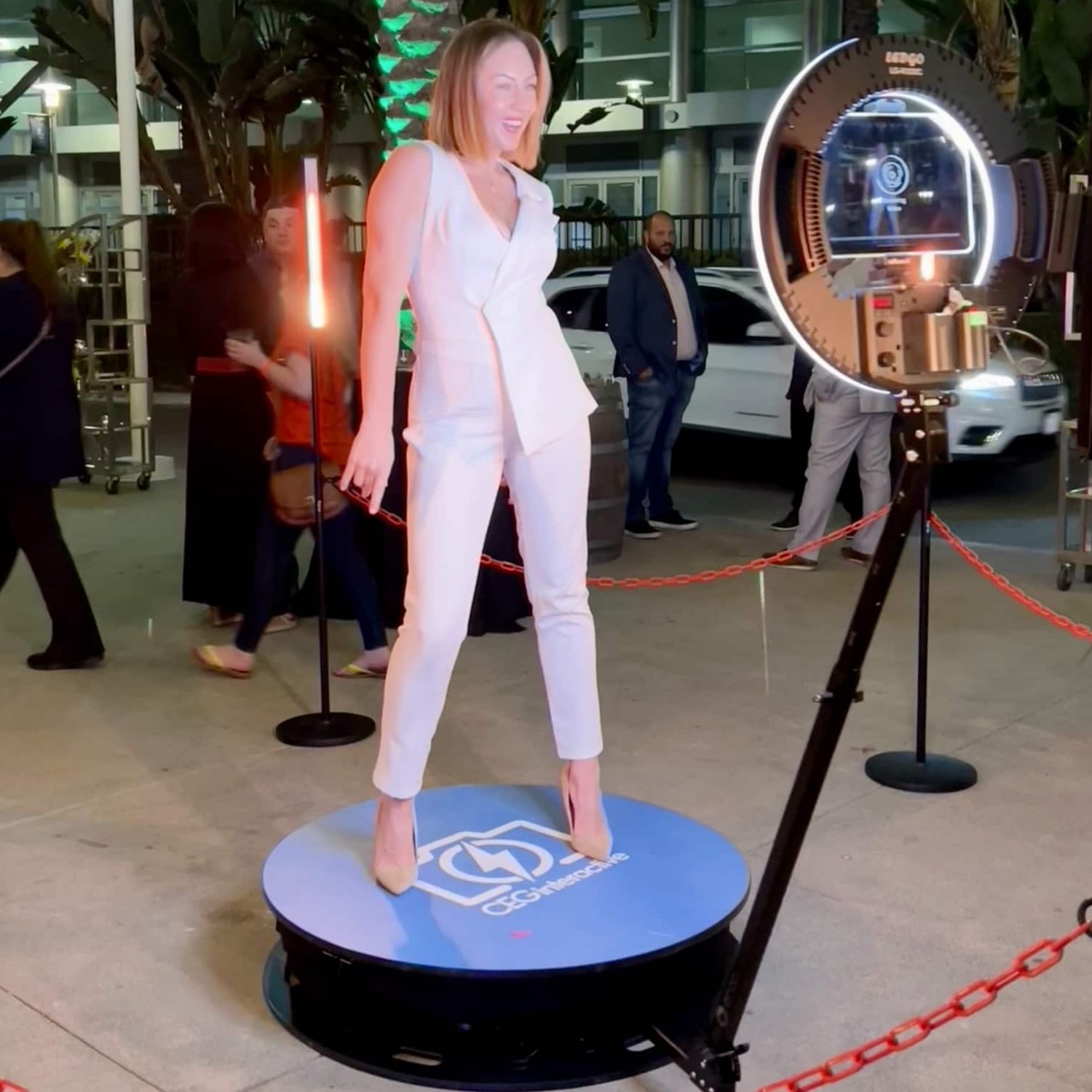 A guest in a white suit standing on a round platform with a logo decal outside a hotel entrance at night while a ring-light camera on an arm circles her, with red chain stanchions around the platform