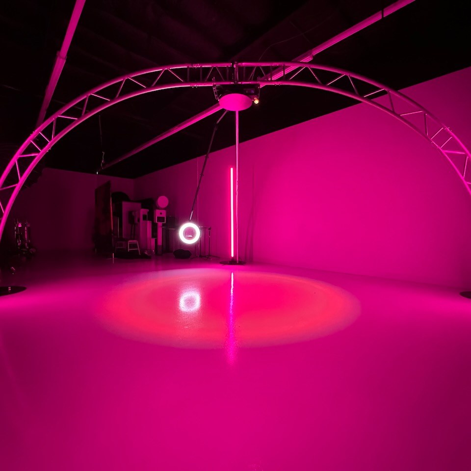 Two curved truss arches crossing overhead in a studio washed in magenta light, with a ring-light camera hanging from the apex on a drop arm above a glossy floor