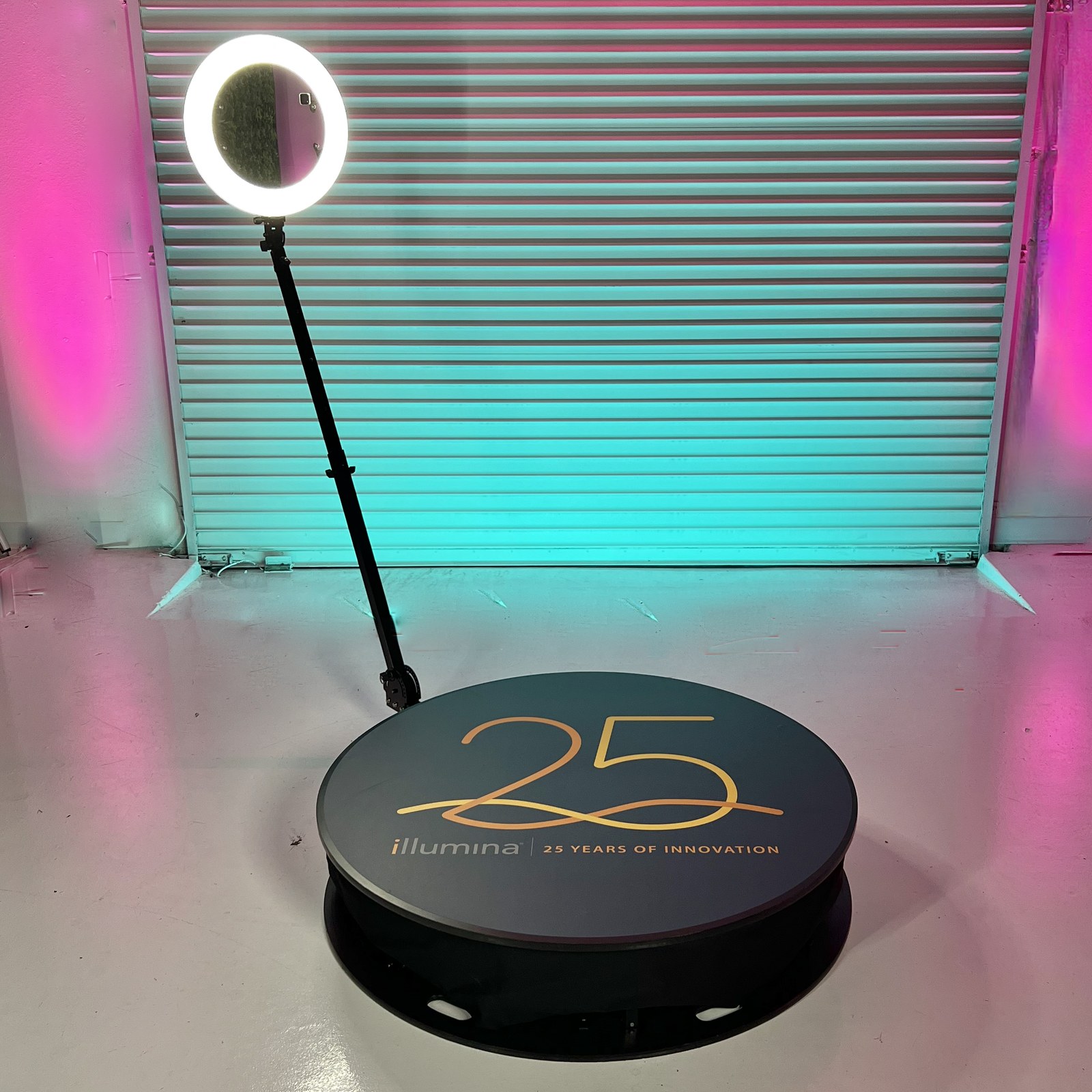 A round platform with a dark decal reading 25 in gold above Illumina, 25 Years of Innovation, with a ring light on the camera arm and a wall washed in teal and pink light