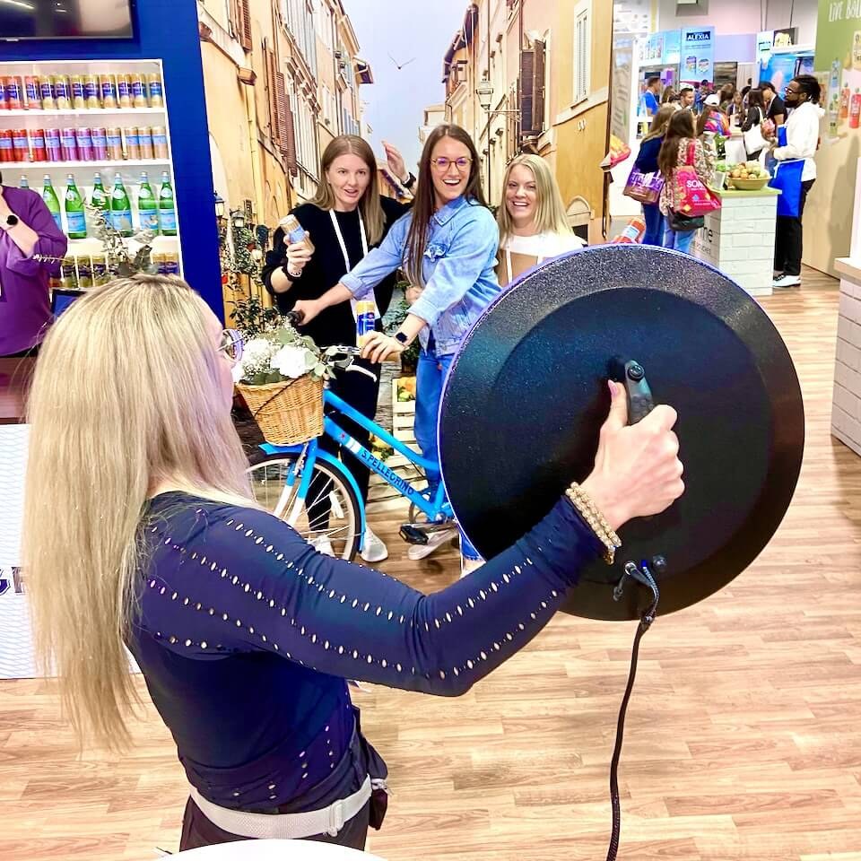 An attendant holds a round handheld photo booth up to three attendees posing with a bicycle at a trade-show booth