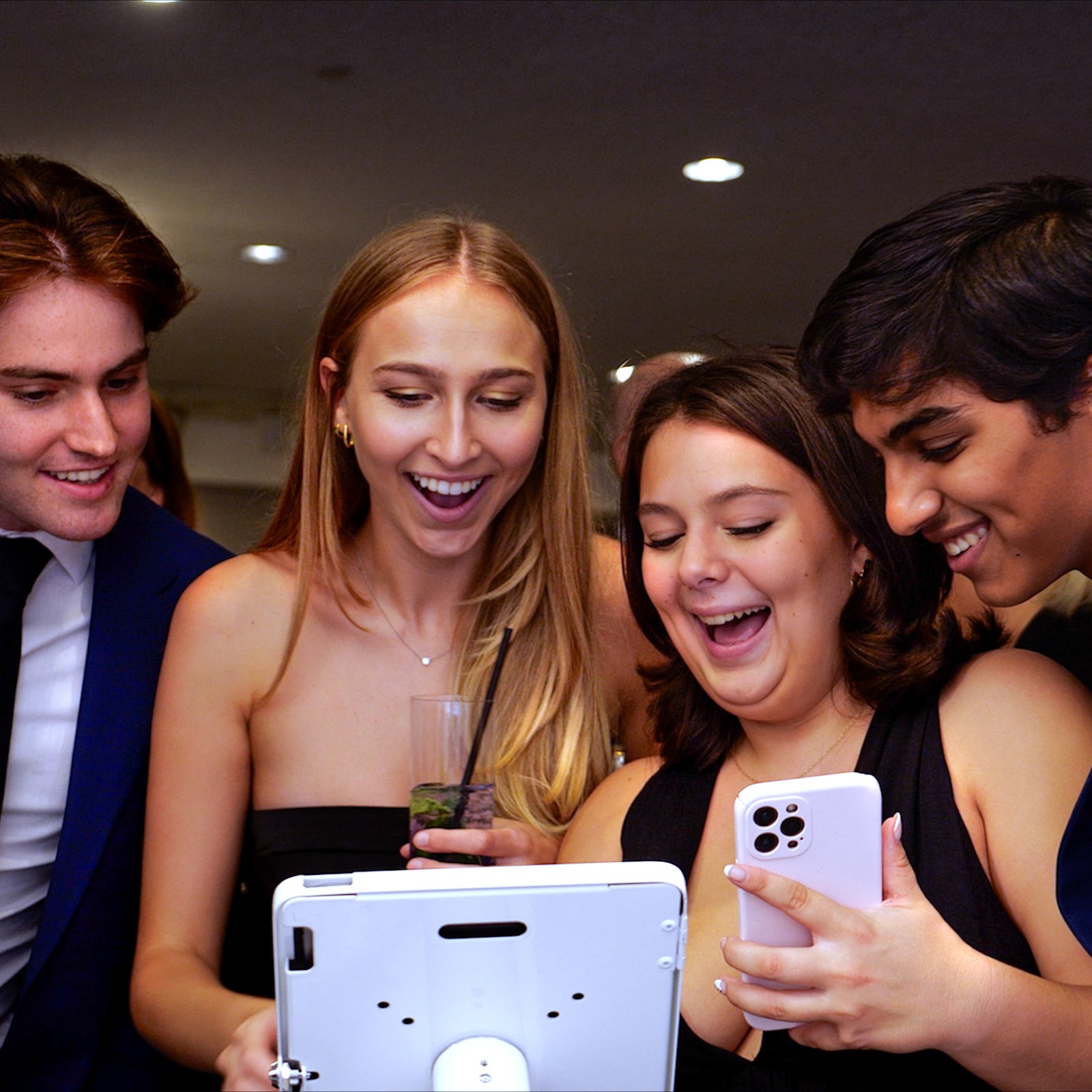 Four guests in evening wear laugh as they watch their clip on a white tablet sharing station, one holding up her phone