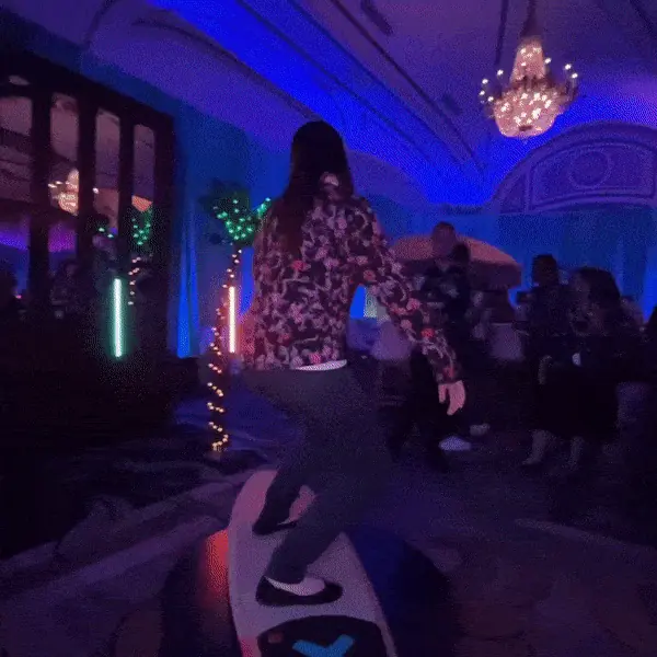 A guest in a patterned jacket rides a surfboard mounted on a round platform in a ballroom lit blue, with LED light tubes and a teal curtain behind her