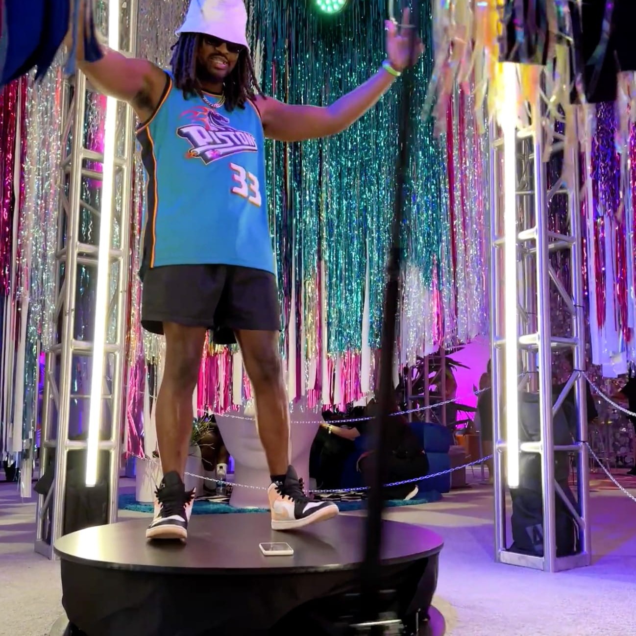 A guest in a bucket hat and a basketball jersey raises his arms on the platform inside a truss frame fitted with white LED tubes, in front of tinsel curtains