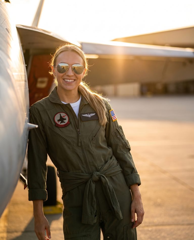 A guest leans against the fuselage of a fighter jet parked on an open tarmac at the last minutes of golden hour.