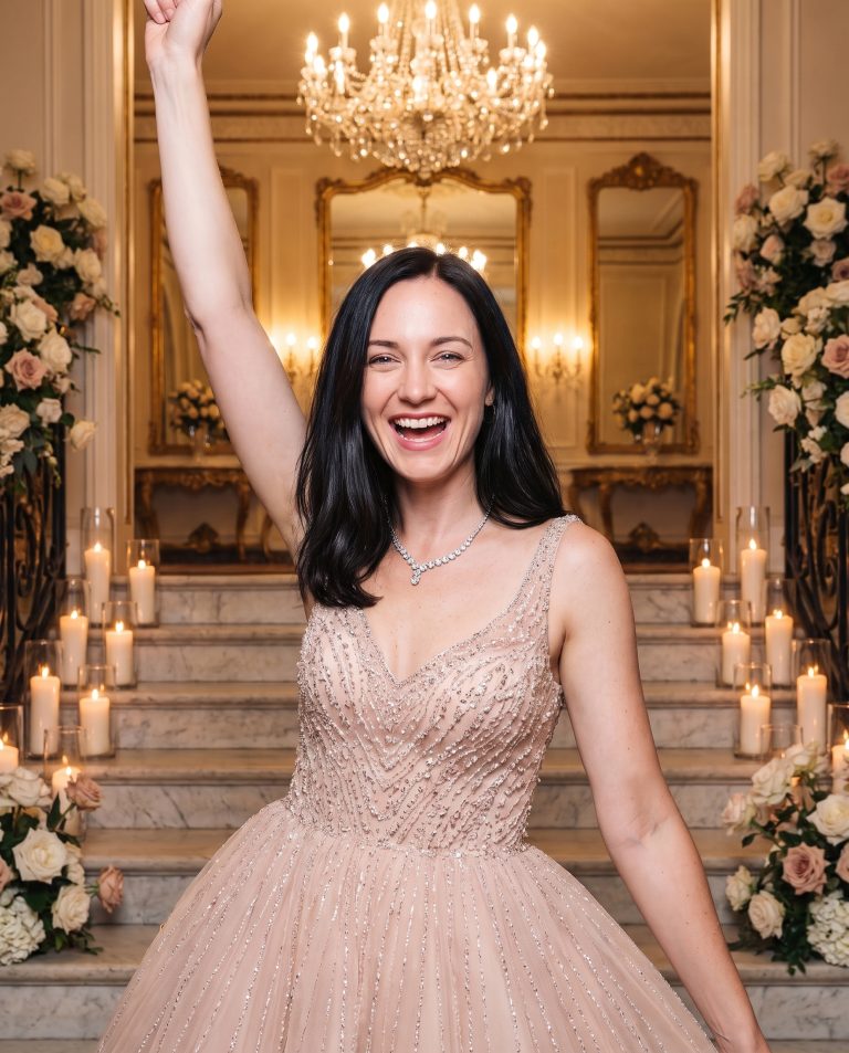 A guest stands at the foot of a sweeping marble staircase in a grand ballroom lit for a celebration.