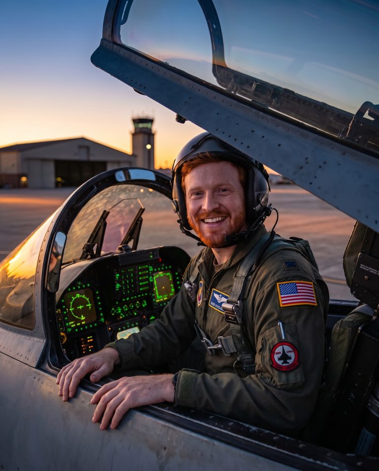 A guest sits in the cockpit of a fighter jet with the canopy raised, hands resting on the canopy rail.