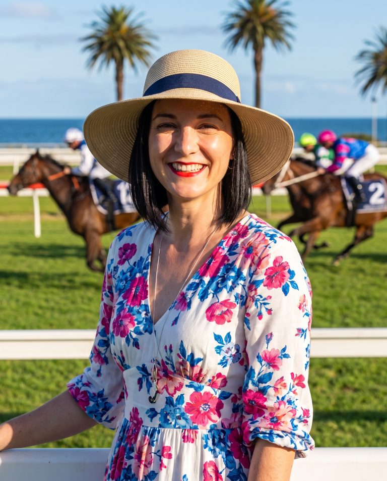 A guest stands trackside at a seaside horse-racing meet on opening day, a manicured green turf course.