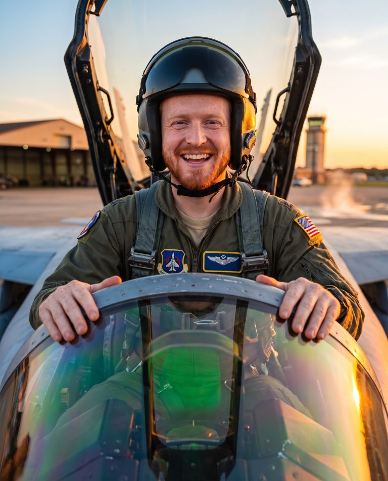 A guest sits in the cockpit of a fighter jet with the canopy raised, hands resting on the canopy rail.