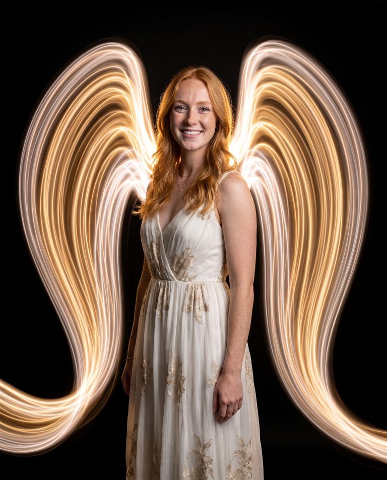 A guest lit sharply in a pitch-black studio with a huge pair of glowing light-painted wings sweeping out behind them.