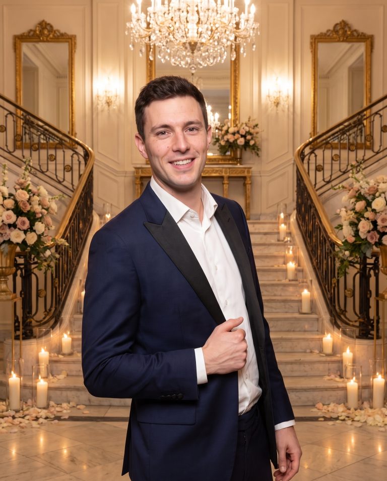 A guest stands at the foot of a sweeping marble staircase in a grand ballroom lit for a celebration.