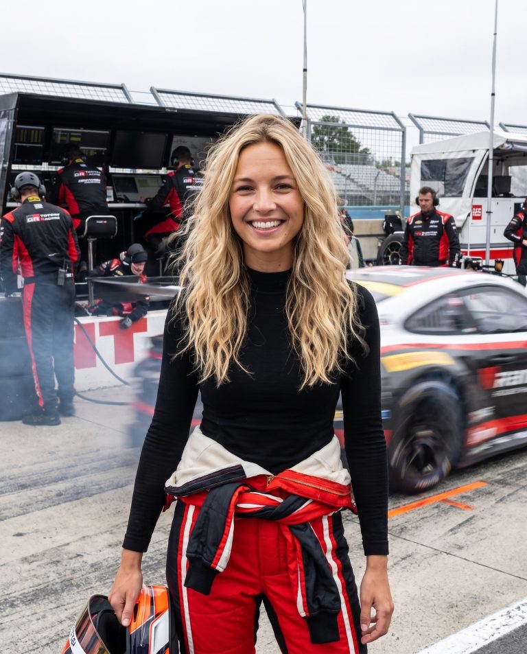 A guest stands in a race pit lane during a session, garage bays glowing behind them with tool walls and monitors.
