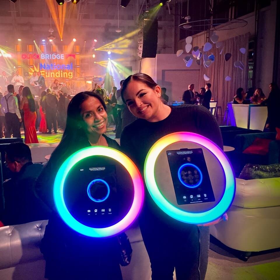 Two attendants holding ring-light tablets among guests at a corporate party, with a lit stage behind them