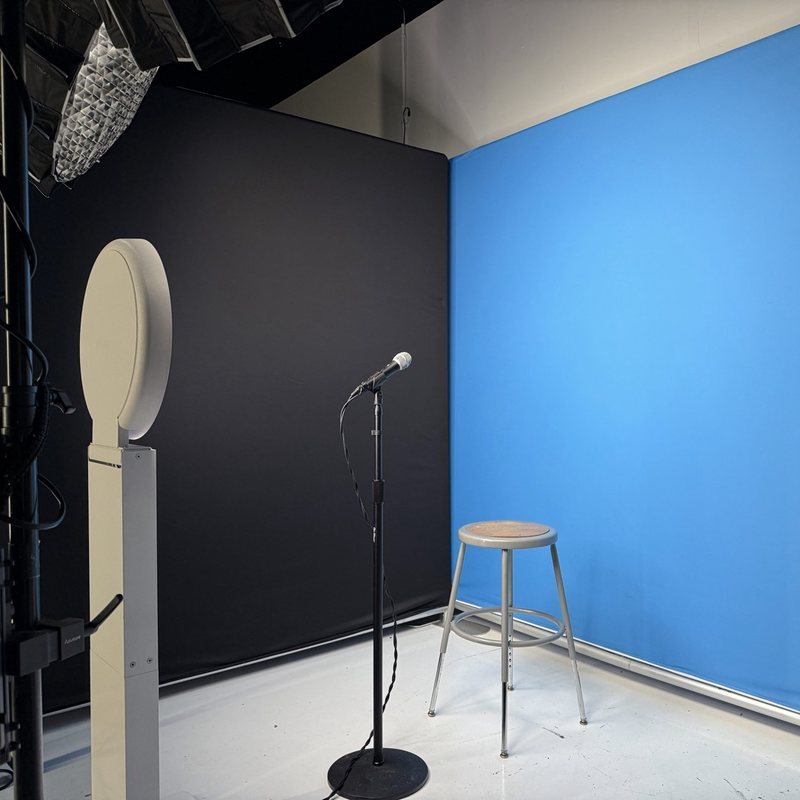 A video interview set with a stool and a microphone in front of a blue backdrop, lit by a softbox beside a white camera stand