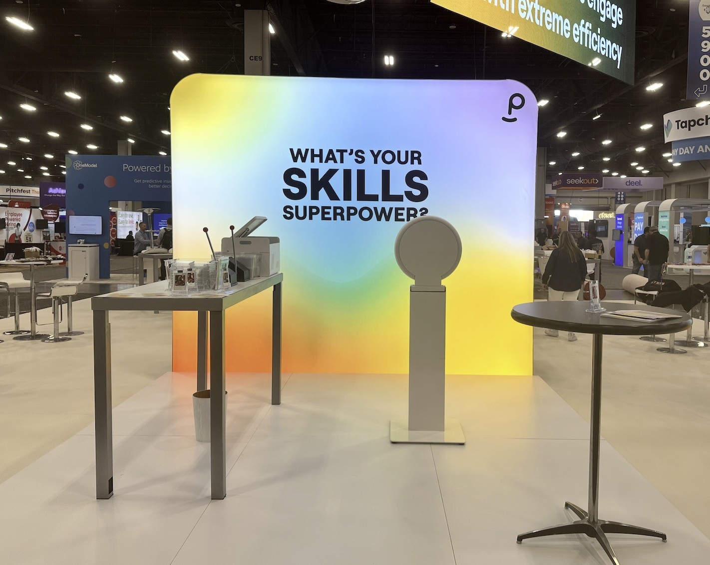 A trade show booth with a rainbow gradient backwall reading WHAT'S YOUR SKILLS SUPERPOWER?, a white touchscreen photo booth on a stand, and a table with a card printer and finished cards in acrylic holders