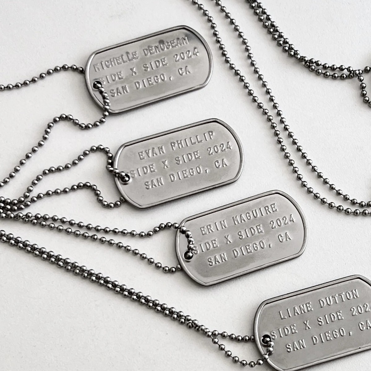 Four embossed stainless steel dog tags on ball chains laid out on a white surface