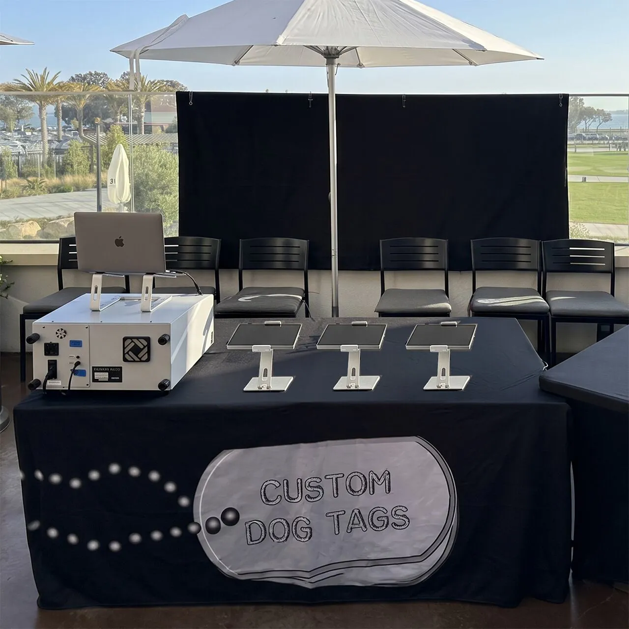 Black-draped table on a covered patio with three landscape tablets on stands, a light-grey embossing machine with a laptop above it, and a printed tag graphic on the table skirt, with a bay and palm trees behind