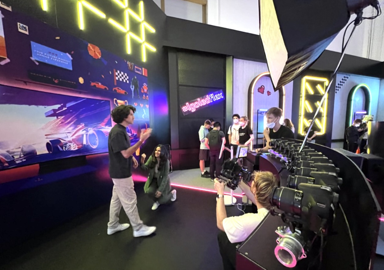 A curved arc of cameras on a show floor photographing two guests in front of a pixel-art wall with a pink neon sign