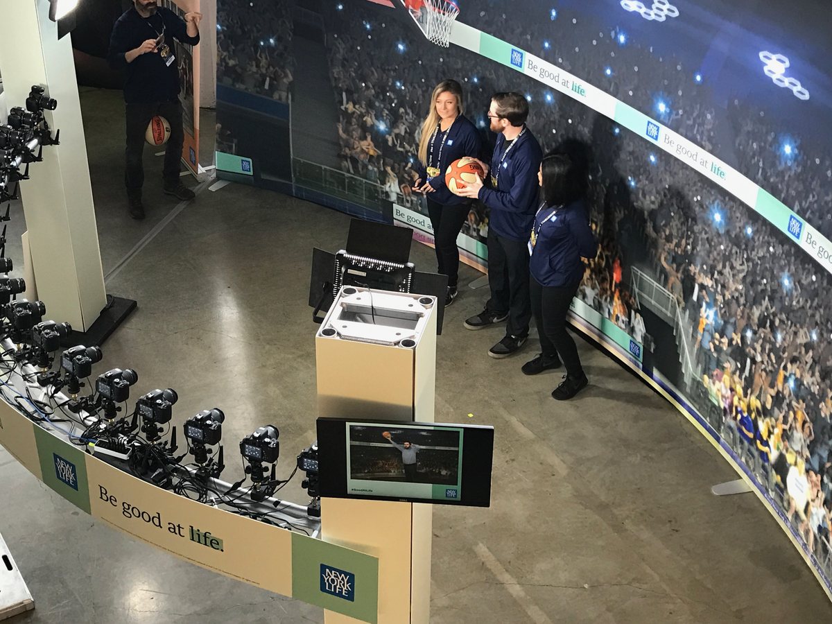 An arc of 18 cameras on white pillars faces three guests holding a basketball in front of a curved arena backdrop