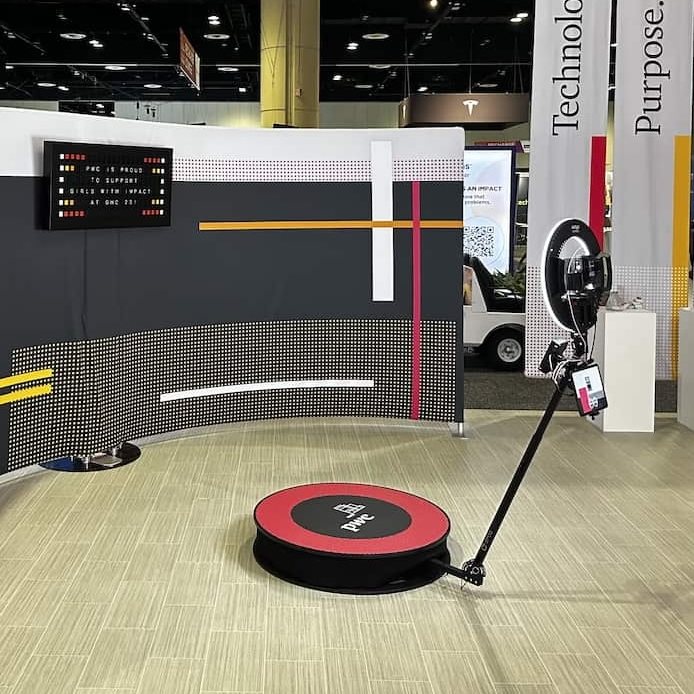 A 360 platform with a PwC decal in front of a curved printed backdrop and a split-flap message board on a convention floor