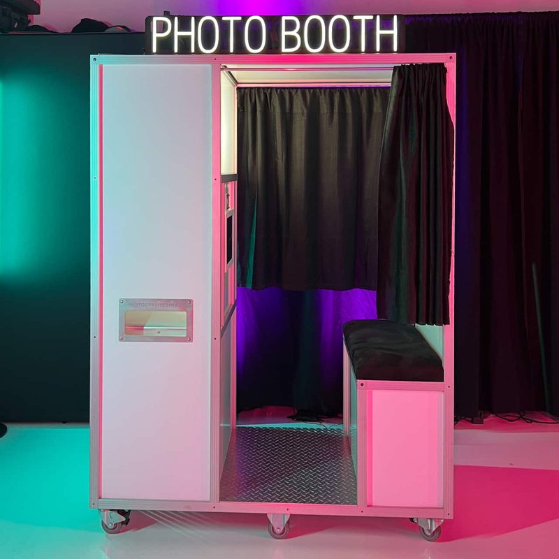 An enclosed photo booth with a black curtain, a bench and a lit Photo Booth sign