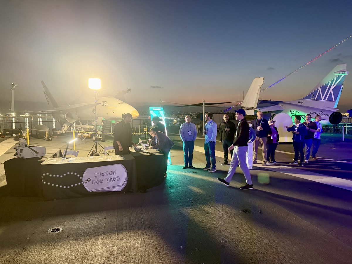 A dog tag station with a Custom Dog Tags sign on an aircraft carrier flight deck at dusk, jets behind it and a line of guests
