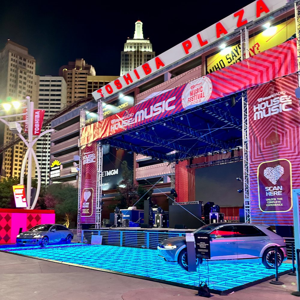 A lit LED floor with two cars parked on it in front of a festival stage at Toshiba Plaza in Las Vegas at night