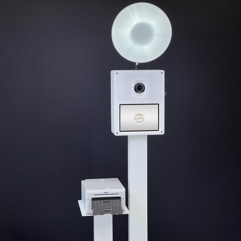 A white photo booth with a round beauty dish light and a touchscreen on a stand, with a photo printer on a shelf, against a dark gray background