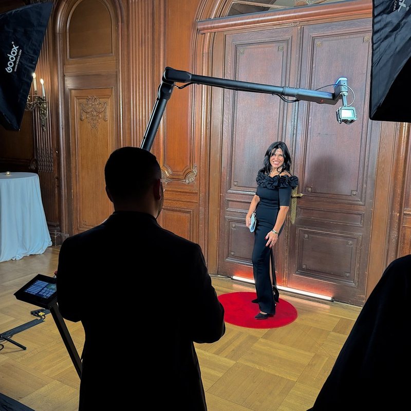 A robotic camera arm films a guest on a red circle in a wood-paneled room while an operator watches