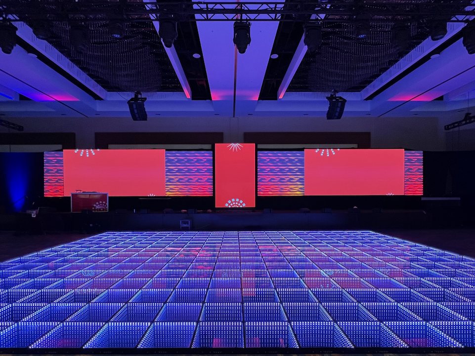 A wide mirror-tile floor glowing blue and violet in front of a general-session stage with red video screens and lighting truss