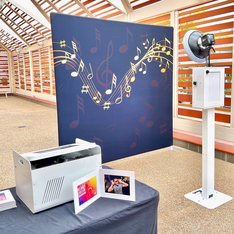 A white photo booth on a stand in front of a navy backdrop with gold music notes, beside a printer and framed 5 by 7 prints