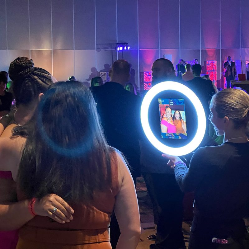 An attendant holds a ring-lit roaming photo booth up to two guests at a dimly lit evening event