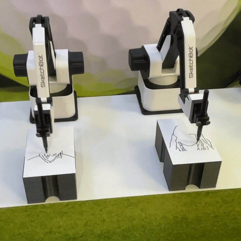 Two small white robot arms draw line portraits on cards