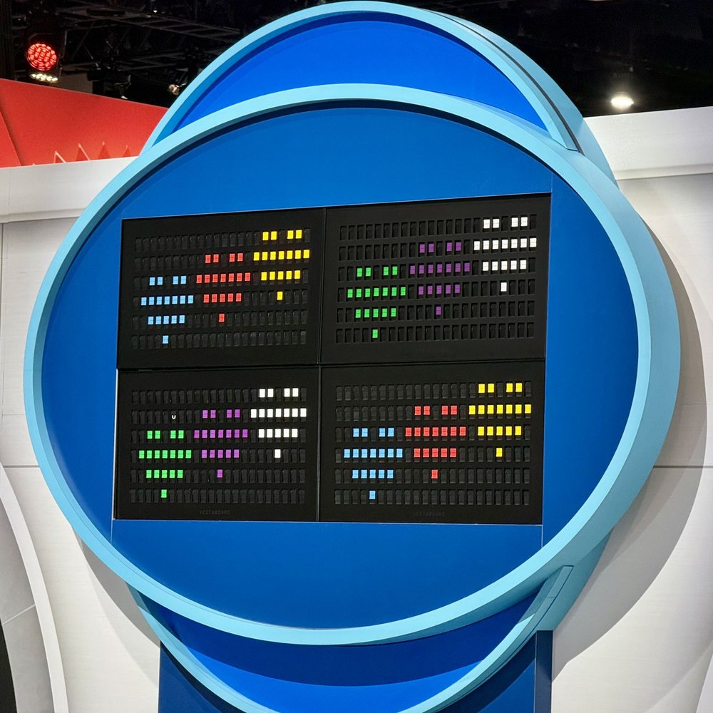 Four split-flap boards showing colored tiles, set into a round blue fabricated wall on a show floor