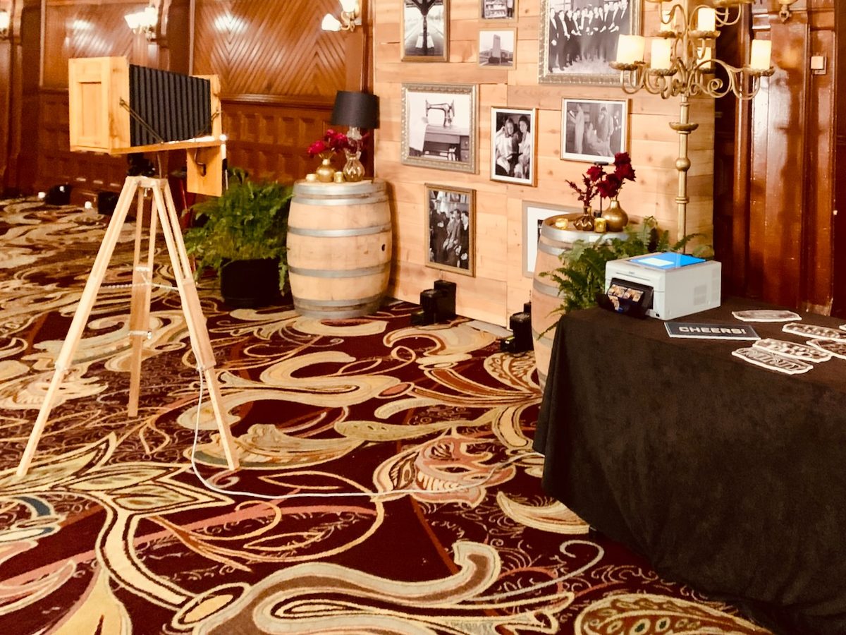 A vintage wood camera booth with a barrel, framed prints and a print table in a wood-paneled Hotel del Coronado ballroom