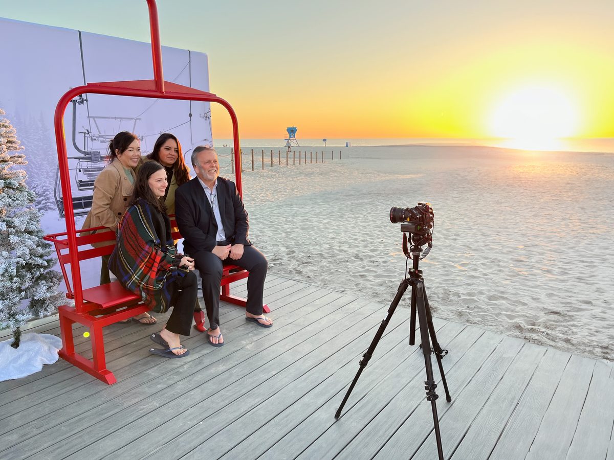 Four guests seated on a red ski lift chair on the Hotel del Coronado beach at sunset with the camera on a tripod in front