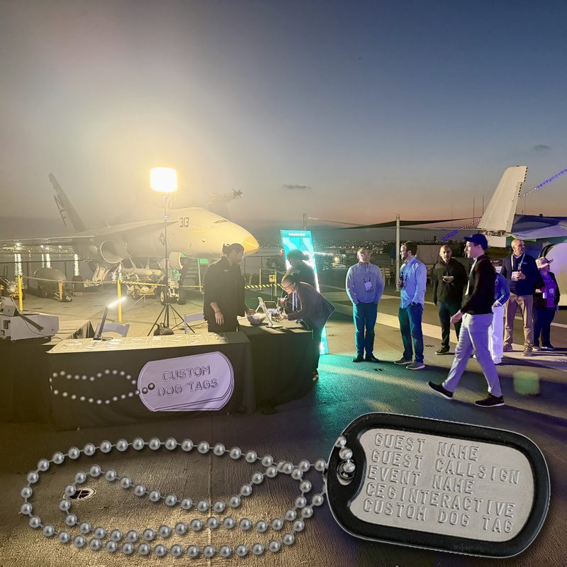 A stamped sample dog tag and its ball chain on the USS Midway flight deck, with guests at a dog tag station behind it at dusk