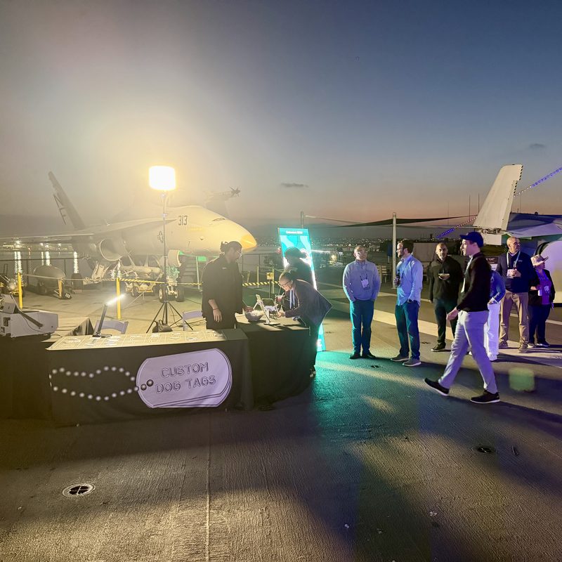 Guests lining up at a dog tag station on the USS Midway flight deck at dusk with jets behind them