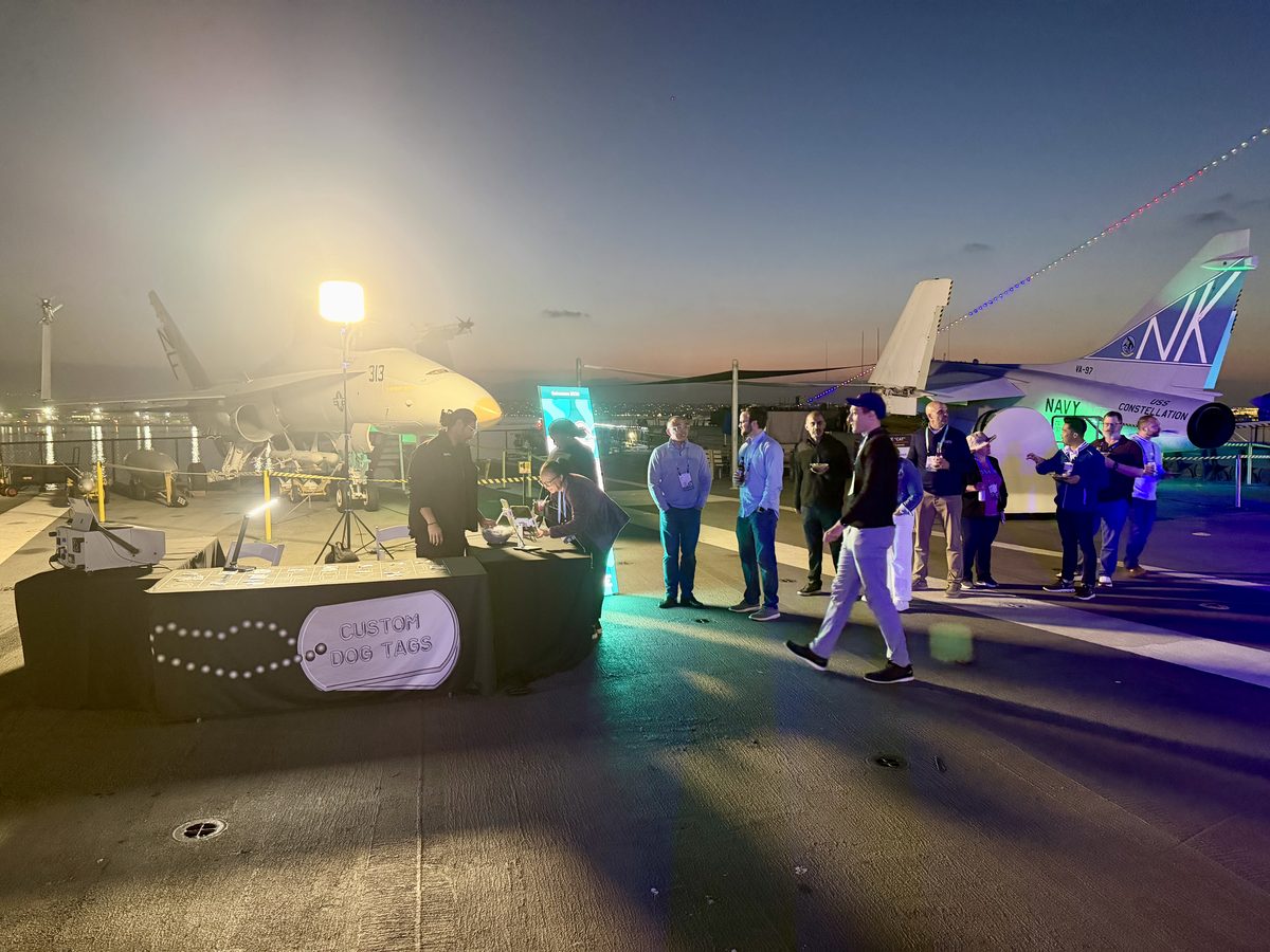 Guests lining up at a dog tag station on the USS Midway flight deck at dusk with jets behind them