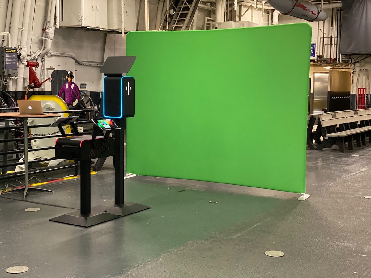 A digital booth and green backdrop set up on the USS Midway hangar deck with the ship structure overhead
