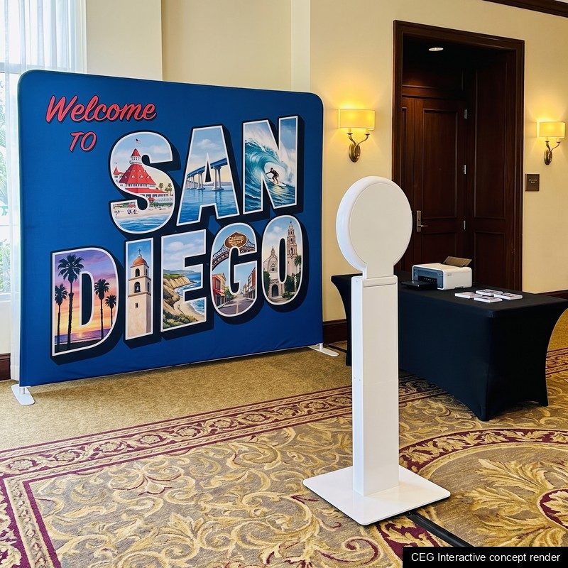 An open-air photo booth stand and a print table in front of a large-letter Welcome to San Diego postcard backdrop in a hotel foyer, concept render