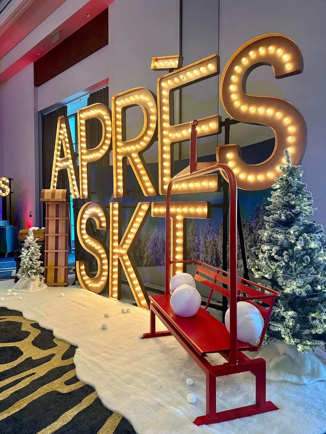 A red ski chairlift bench with plush snowballs beside a large lit APRES SKI marquee and a flocked Christmas tree
