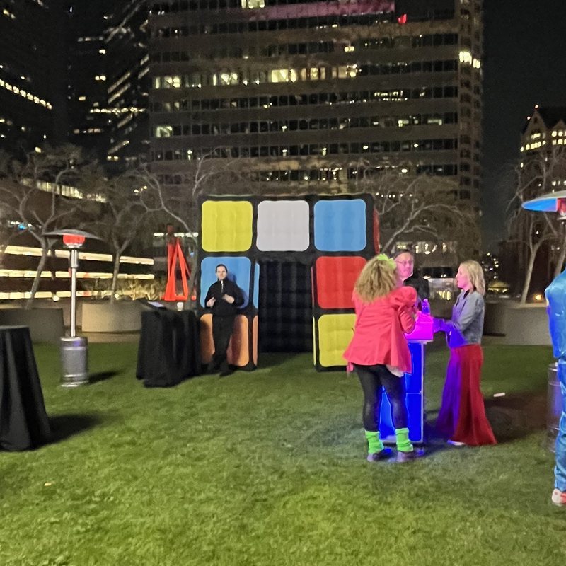 A giant lit cube photo booth on a rooftop lawn at night, with guests in bright costumes beside a glowing cocktail table